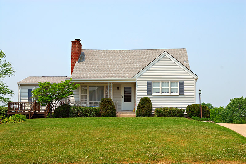 USDA Loan 1 A single-story house with light gray siding, dark shutters, and a brick chimney, sits on a neatly trimmed lawn with bushes and a wooden deck on the left side.