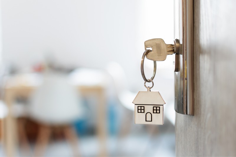 A key with a house-shaped keychain is inserted into a door lock, with a blurred interior room visible in the background.