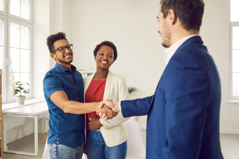 A smiling man in a blue shirt shakes hands with a man in a suit while a woman in a white blazer stands beside them, all in a bright office setting.