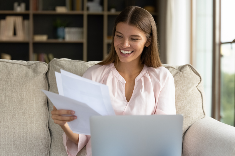 Bank Statement Program 1 A smiling woman sits on a couch holding papers in one hand while looking at a laptop, appearing happy and relaxed in a cozy, well-lit living room with bookshelves in the background.