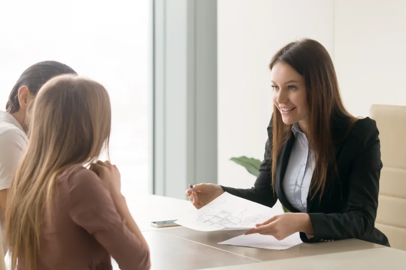 A smiling woman in a business suit sits at a desk, holding papers and a pen, talking to two people seated across from her in a bright office setting.