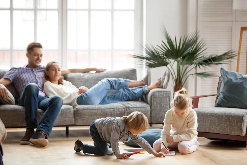A man and woman relax together on a sofa while two young children draw and color on the floor in a bright, cozy living room with large windows and a potted plant.