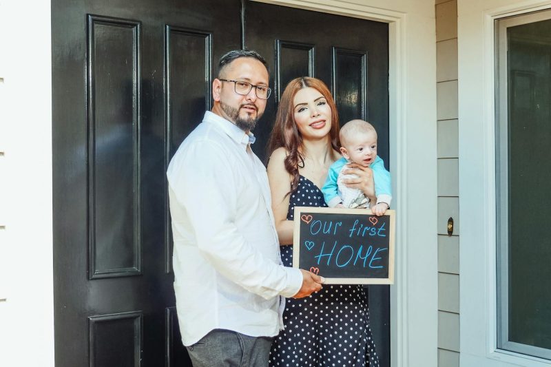 Blog 1 Young family with baby holding a sign Our First Home at door